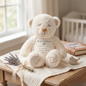 Può includere: Orsacchiotto color crema con naso a forma di cuore e occhi chiusi. L'orso ha un pannello personalizzato con il nome "Baby Anderson" e il testo "CARRIED FOR A moment LOVED FOR A lifetime." Il piede dell'orso ha l'anno "2026" e un cuore d'oro.