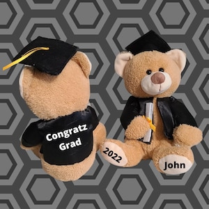 Könnte beinhalten: Ein brauner Teddybär, der eine schwarze Graduationskappe und -robe trägt. Der Bär hält ein Diplom in seiner Pfote. Die Pfote des Bären trägt das Jahr 2022. Die andere Pfote des Bären trägt den Namen John. Der Rücken des Bären trägt den Text "Congrats Grad".