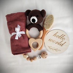May include: A baby gift set featuring a brown crocheted bear rattle, a wooden heart-shaped teether, a wooden brush, a wooden clip-on teether, and a wooden sign that reads "hello world".
