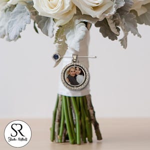 May include: A silver pendant with a photo of two people, surrounded by clear crystals, attached to a bouquet of white roses and greenery. A small blue gem is pinned to the bouquet. The bouquet is wrapped in white ribbon.