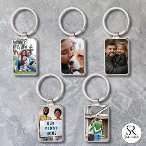 Puede incluir: Llaveros rectangulares plateados con fotos personalizadas. Los llaveros presentan retratos familiares, un perro y una pareja con un letrero que dice "NUESTRA PRIMERA CASA". Los llaveros están sujetos a llaveros plateados.