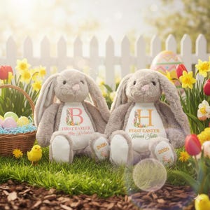 May include: Two plush bunny rabbits with long ears, sitting on grass in front of a white picket fence. Each bunny has a personalized chest design with a letter, the words "First Easter", and a name. The year "2026" is on the foot.