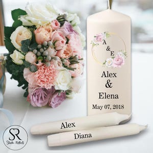 May include: A cream-coloured pillar candle with floral accents and the names "Alex & Elena" and the date "May 07, 2018". Two taper candles are labelled "Alex" and "Diana". A bouquet of pink, peach, and white flowers is in the background.