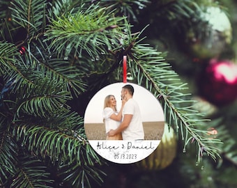 Könnte beinhalten: Ein runder Weihnachtsschmuck mit einem Foto eines Paares. Der Schmuck trägt die Namen "Alice & Daniel" und das Datum "05.13.2023". Er hängt an einem roten Band vor dem Hintergrund eines Weihnachtsbaums.