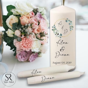 May include: A cream-colored pillar candle with a gold floral wreath design and the names "Alan & Diana" printed in black. Two taper candles with the names "Alan" and "Diana" are also present. A bouquet of pink, white, and purple flowers is in the background.