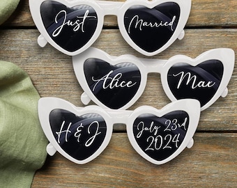 May include: Four pairs of white heart-shaped sunglasses with black lenses. The sunglasses are decorated with white text, including "Just Married", "Alice", "Max", "H & J", and "July 23rd 2024".