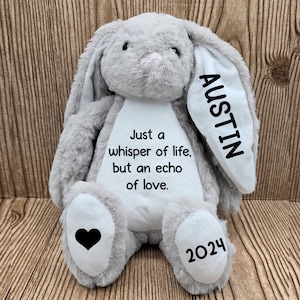 Op de afbeelding: Een grijze pluche konijn met lange oren, geborduurd met de naam "Austin" op één oor. Het konijn heeft de tekst "Just a whisper of life, but an echo of love" op de borst, een zwart hart en het jaartal "2024" op de voet.