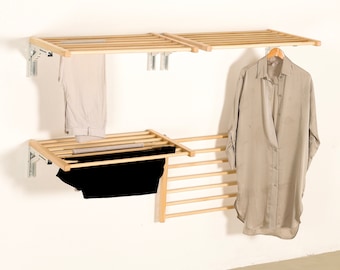 Puede incluir: Un tendedero de pared de madera con múltiples listones horizontales y soportes metálicos ajustables. La unidad incluye dos estantes superiores y una sección inferior, con pantalones claros, una prenda negra y una camisa beige colgadas.
