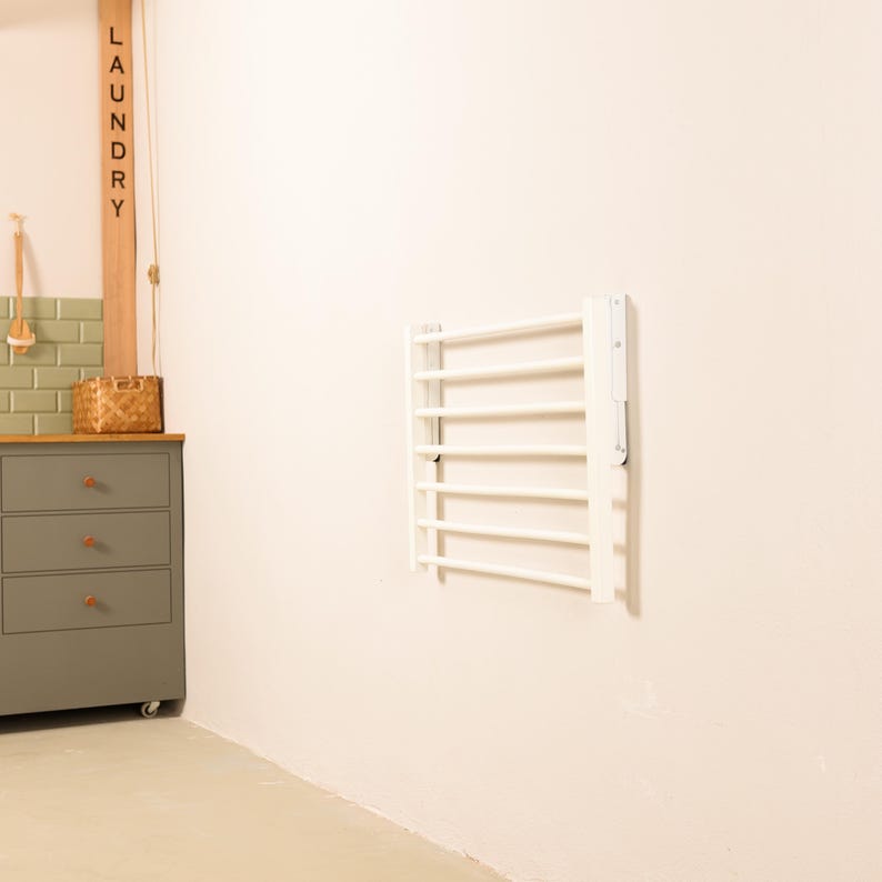 May include: White wall-mounted folding clothes drying rack with six bars. The rack is folded up and mounted on a white wall. The word "LAUNDRY" is written on a wooden sign to the left of the rack.