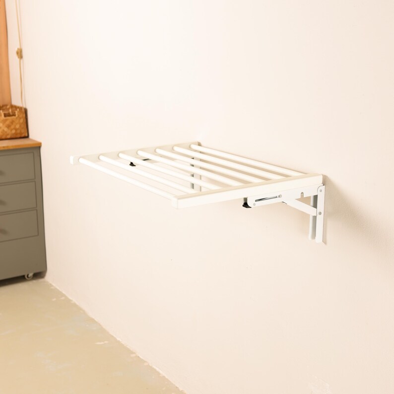 May include: White wall-mounted folding clothes drying rack with six metal bars. The rack is folded up and mounted on a white wall.