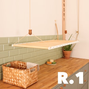 May include: A white ceiling-mounted drying rack with wooden dowels hangs above a wooden countertop. The countertop is in a laundry room with green cabinets and a woven basket.