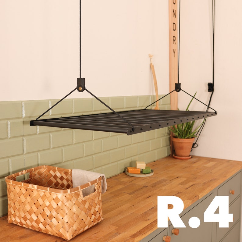 May include: A black metal hanging drying rack with a wooden sign that says "Laundry" hanging above it. The rack is suspended from the ceiling by black cords. The rack is in a room with green tile walls and a wooden countertop. There is a wicker basket and some soap bars on the countertop.