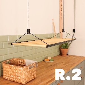 May include: A black metal and wood hanging clothes drying rack with a wooden slat design. The rack is suspended from the ceiling and is hanging over a wooden countertop with a woven basket and green cabinets. The text "R.2" is visible on the cabinet.