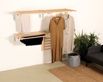 May include: A wall-mounted wooden drying rack system with multiple slatted shelves and folding arms. The unit features light-colored wood slats, metal brackets, and hangers holding various garments including a brown dress and a light gray shirt.