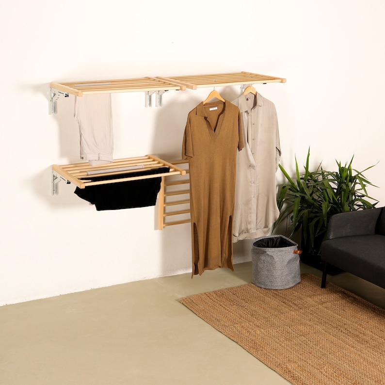 May include: A set of three wooden wall-mounted shelves with clothes hanging on them. The shelves are made of light wood and have a simple design. There is a gray laundry basket on the floor below the shelves.