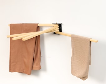 May include: A wall-mounted wooden clothes drying rack with two brown garments hanging from it. The rack is made of four wooden dowels that are connected by a black metal bracket.
