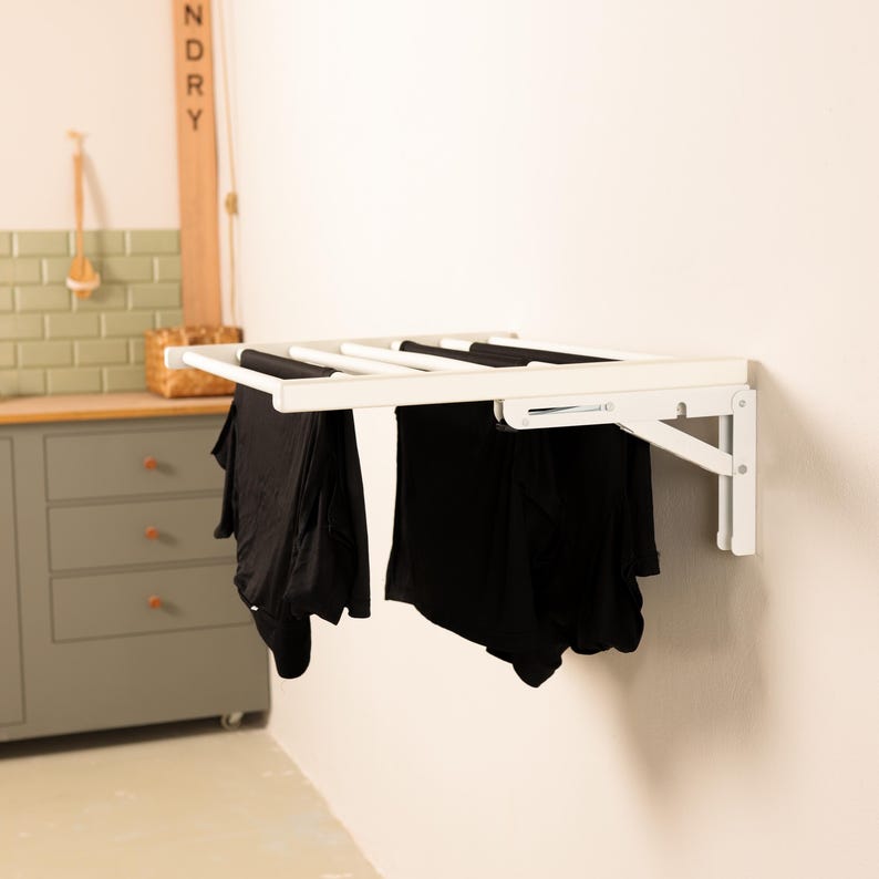 May include: White wall-mounted folding clothes drying rack with four bars. The rack is folded up and has black clothing hanging from it.