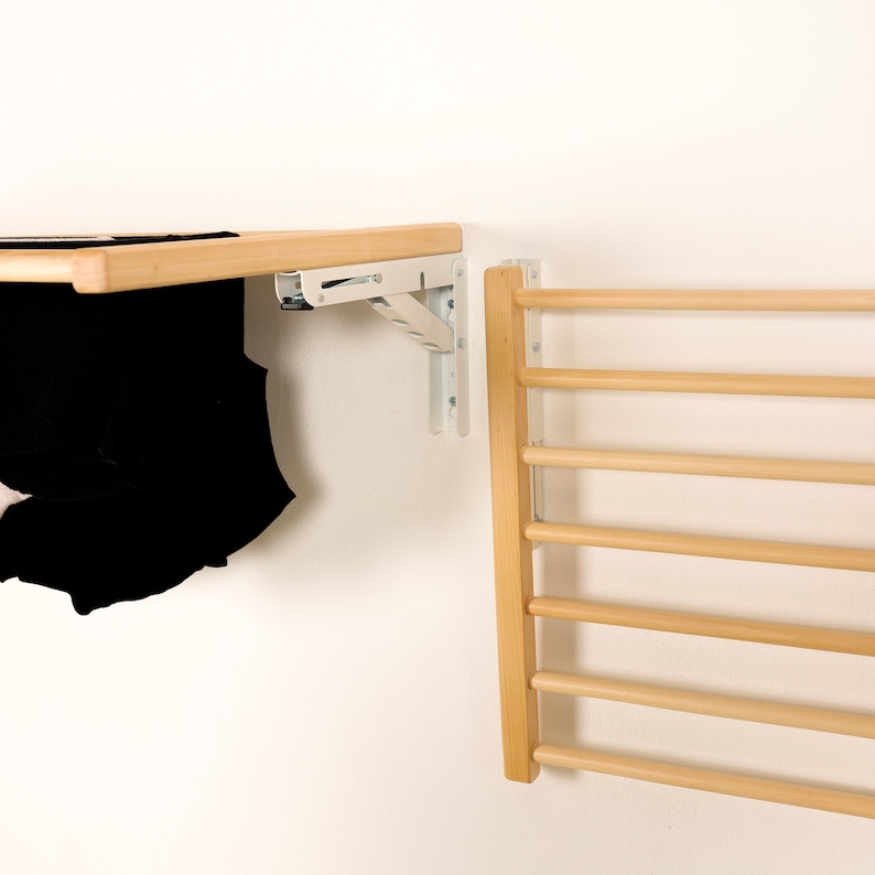 May include: A wooden wall-mounted drying rack with six horizontal bars. The rack is made of natural wood and has a light brown finish. A black t-shirt is hanging on a wooden shelf above the rack.