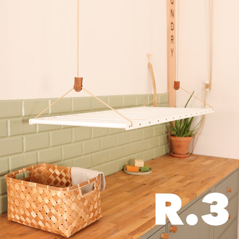 May include: White hanging clothes drying rack with a wooden sign that says "Laundry" on the wall. The rack is suspended from the ceiling with rope and leather straps. A woven basket and a potted plant are on the counter below the rack.
