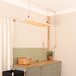 May include: A ceiling-mounted wooden laundry drying rack with white metal side rails, suspended by a pulley system with ropes. The rack features horizontal wooden slats and is positioned above a wooden countertop with a green tiled backsplash.