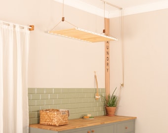 May include: A ceiling-mounted wooden laundry drying rack with white metal side rails, suspended by a pulley system with ropes. The rack features horizontal wooden slats and is positioned above a wooden countertop with a green tiled backsplash.