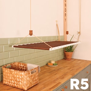 May include: A ceiling-mounted laundry drying rack featuring dark brown wooden dowels held by white metal side frames. The rack is suspended by light-colored ropes attached to leather tabs. A woven basket sits on a wooden countertop below.