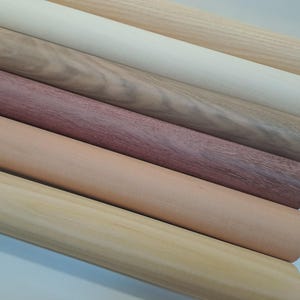 Könnte beinhalten: Eine Sammlung von Holzdübeln in verschiedenen Farben und Maserungen. Die Dübel reichen von hellbeige bis tiefem Rotbraun und zeigen die natürlichen Variationen des Holzes. Die glatten, zylindrischen Formen sind diagonal angeordnet und heben ihre unterschiedlichen Texturen hervor.