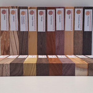 Könnte beinhalten: Eine Präsentation verschiedener Holzproben, jede mit einzigartiger Maserung und Farbe. Die Proben sind mit der Holzart beschriftet, darunter Zebrano, Amaranth und Osage Orange. Die Holzblöcke sind in einer Reihe angeordnet und zeigen ihre natürlichen Texturen.
