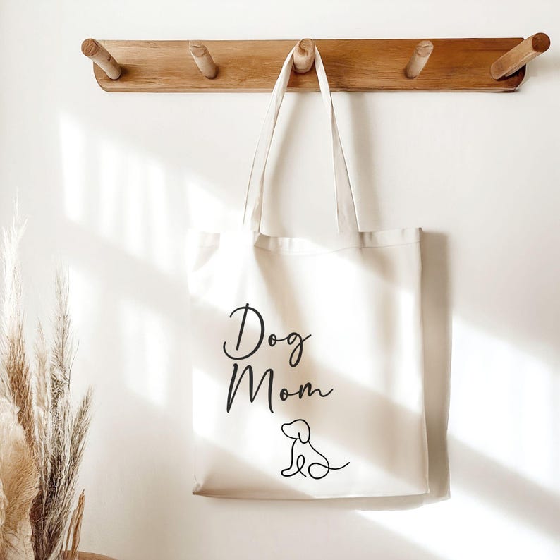 May include: A cream-coloured tote bag featuring the words "Dog Mom" in elegant script, with a simple dog illustration. The bag is suspended from a wooden coat rack on a white wall.