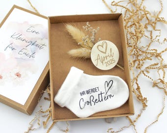 Könnte beinhalten: Eine kleine Geschenkbox mit einer weißen Socke, auf der "Ihr werdet Großeltern" steht, und einem Holz-Kreis mit einem Herz und dem Text "April 2025".