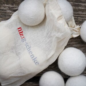 Op de afbeelding: Een verzameling witte wollen droogballen die uit een natuurlijke canvas tas komen. De tas heeft de tekst "filz:g-schenke" in rood en grijs. De ballen zijn gerangschikt op een verweerd houten oppervlak.