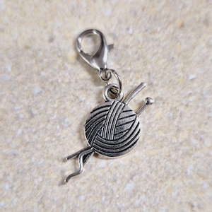 Könnte beinhalten: Ein silberfarbener Metallanhänger in Form eines runden Wollknäuels mit zwei gekreuzten Stricknadeln, befestigt an einem kleinen Karabinerverschluss. Der Anhänger hat ein dunkles, oxidiertes Finish in den Vertiefungen, um die Struktur zu betonen.