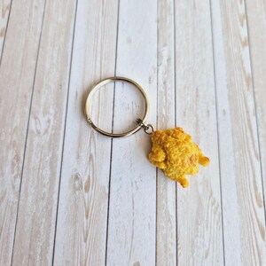 Könnte beinhalten: Ein goldfarbener Schlüsselanhänger mit einem kleinen, strukturierten, gelben Brathähnchen-Nugget-Anhänger. Der Schlüsselanhänger hat einen kreisförmigen Ring und eine kleine Kette, die mit dem Anhänger verbunden ist. Der Hintergrund ist ein helles Holzmaserungsmuster.