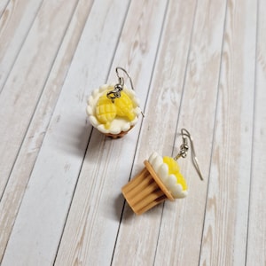 Könnte beinhalten: Ein Paar ausgefallene Ohrringe mit Miniatur-Desserts. Ein Ohrring ist ein Kuchen mit weißem Boden und gelber Füllung. Der andere Ohrring ist ein Cupcake mit gelber Spitze und weißem Zuckerguss. Beide haben silberfarbene Ohrhaken.