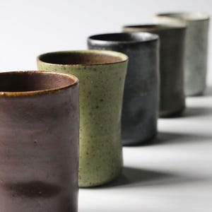 May include: A row of five ceramic tumblers in varying earth tones. The colors include brown, olive green, charcoal, dark grey, and off-white. The cylindrical cups have a slightly textured surface and a simple, minimalist design.