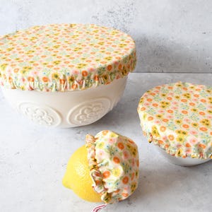Könnte beinhalten: Weiße Keramikschalen mit floralen Stoffschalenabdeckungen. Die Abdeckungen haben ein Muster aus kleinen gelben, orangefarbenen und rosa Blumen auf cremefarbenem Hintergrund. Eine Abdeckung ist auf einer Zitrone. Für die Aufbewahrung von Lebensmitteln.