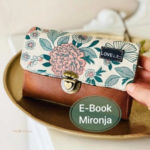 Può includere: Un portafoglio in pelle marrone con una patta in tessuto a stampa floreale. La patta presenta un motivo floreale verde e rosa con il testo "LOVE it" in nero. Il portafoglio ha una chiusura in ottone con un dettaglio a forma di cuore.