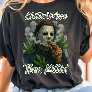 May include: A black t-shirt with a graphic of a man with a white mask smoking a joint. The text "Chillin' More Than Killin'" is printed on the shirt.