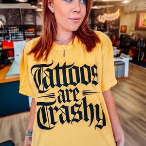 May include: A yellow t-shirt with the phrase "Tattoos are Trashy" in black gothic lettering. The shirt is worn by a person with red hair and is set in a tattoo parlor.