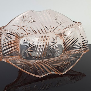Könnte beinhalten: Eine blassrosa Schale aus Pressglas mit einem geometrischen Stern- und Fächermuster. Die Schale hat einen rechteckigen Boden mit ausgestellten, gewellten Seiten und eine klare, reflektierende Oberfläche, die die detaillierten Glasprägungen betont.