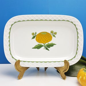 May include: A white rectangular serving dish with a green and white scalloped border. The dish features a yellow flower with green leaves and stems in the centre.