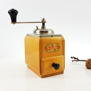 Peut inclure: Un moulin à café vintage en bois avec une poignée noire et un dessus argenté. Le moulin possède un tiroir pour recueillir le café moulu et une étiquette portant l'inscription "Lehmann" en lettres rouges.