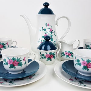 Op de afbeelding: Een wit theeservies met een bloemenpatroon in roze en groen. De set bevat een theepot, suikerpot, roomkan en vier theekopjes met schoteltjes. De schoteltjes zijn tealblauw.