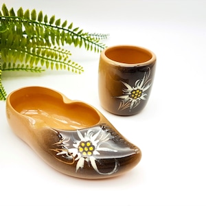Op de afbeelding: Een set van twee bruine keramische bekers met een wit edelweiss-bloemdessin. De bekers zijn gevormd als een traditionele Duitse schoen.