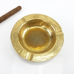May include: A round, gold-coloured brass ashtray with a slightly raised rim and a small indentation in the centre.