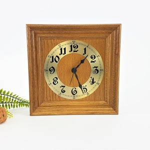 May include: Square wooden clock with a light brown frame. The clock face is gold with black numerals and hands. The clock is set against a white background with a fern and a walnut.