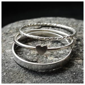 Puede incluir: Tres anillos de plata apilados uno encima del otro. El anillo superior es una banda fina con un dije en forma de corazón. El anillo del medio es una banda fina con un diseño trenzado. El anillo inferior es una banda más gruesa.