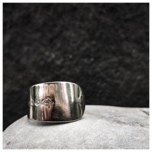 May include: A wide, silver ring with a polished surface. The ring has a diamond-shaped design and other markings. The ring is displayed on a grey stone surface against a dark background.