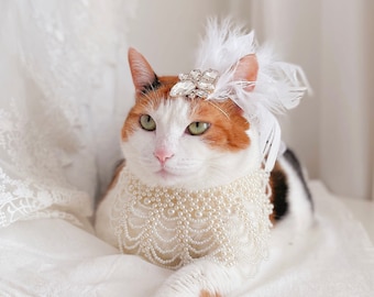 May include: A calico cat wearing a white pearl necklace and a white feathered headband with a rhinestone brooch. The cat is lying on a white fabric background.