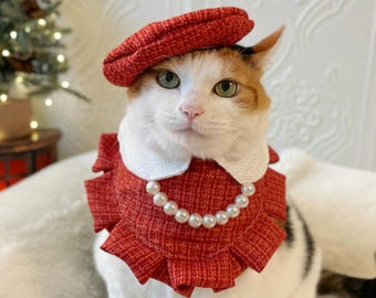 May include: A cat wearing a red beret and a matching red ruffled collar with a pearl necklace. The cat has white and orange fur and green eyes. The collar has a white lace trim.